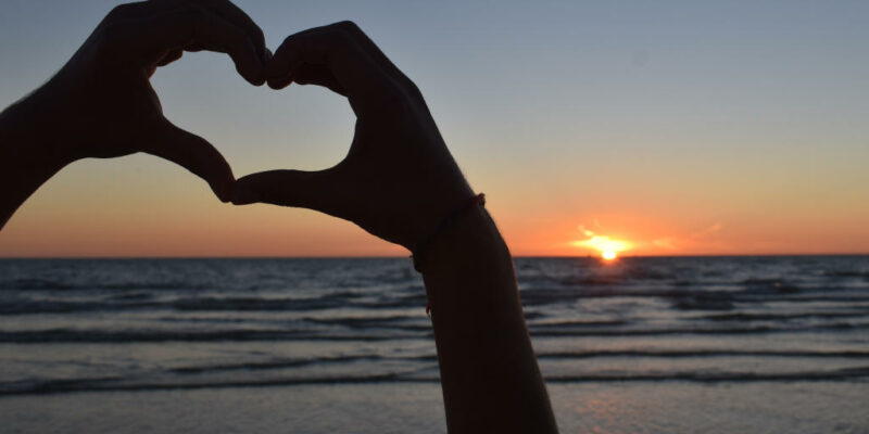 Hände die ein Herz formen mit dem Meer und dem Sonnenuntergang als Hintergrund